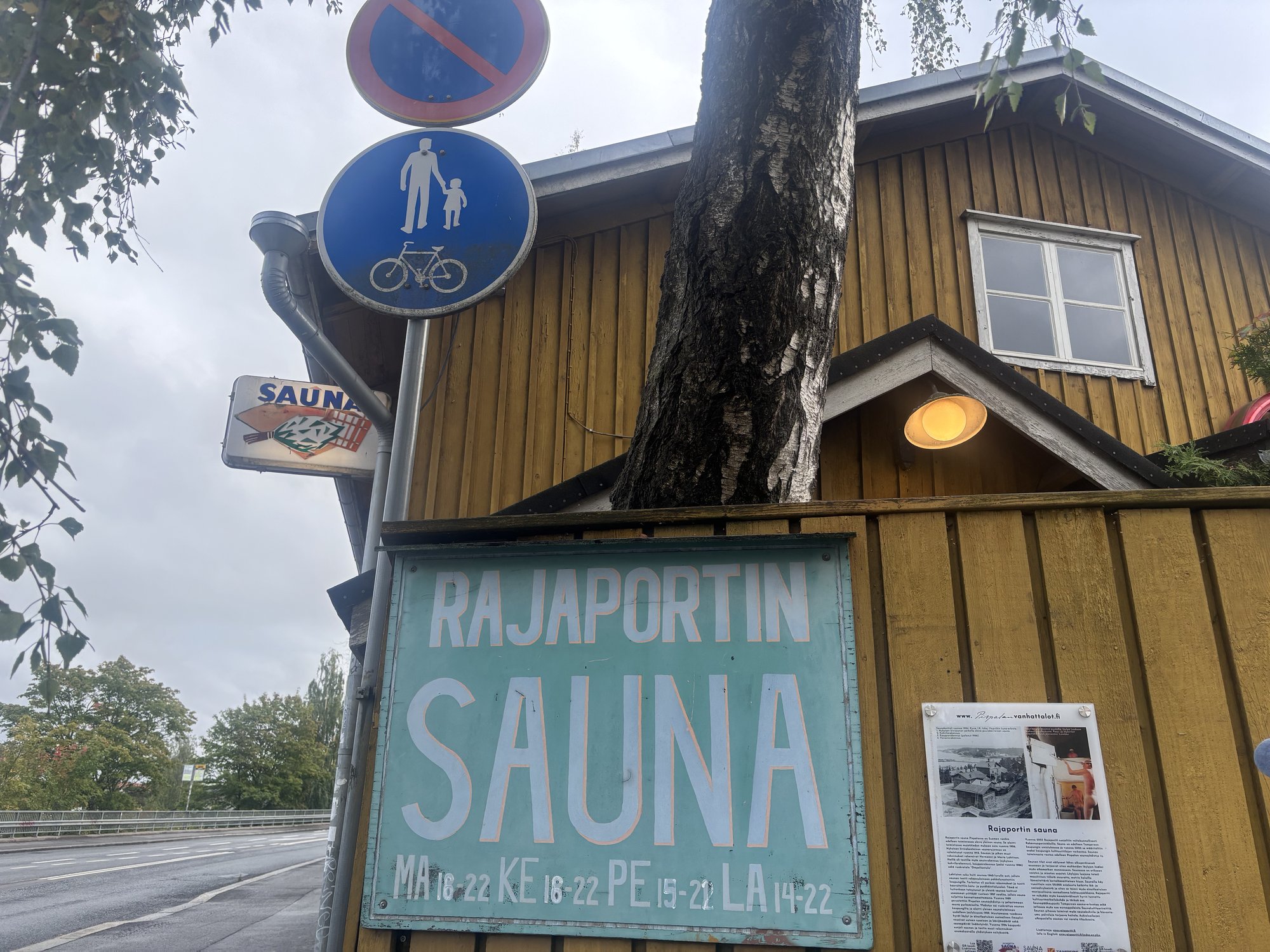 「RAJAPORTIN SAUNA」の看板と、その下に営業時間の表示、隣に歴史説明のプレートが見える
